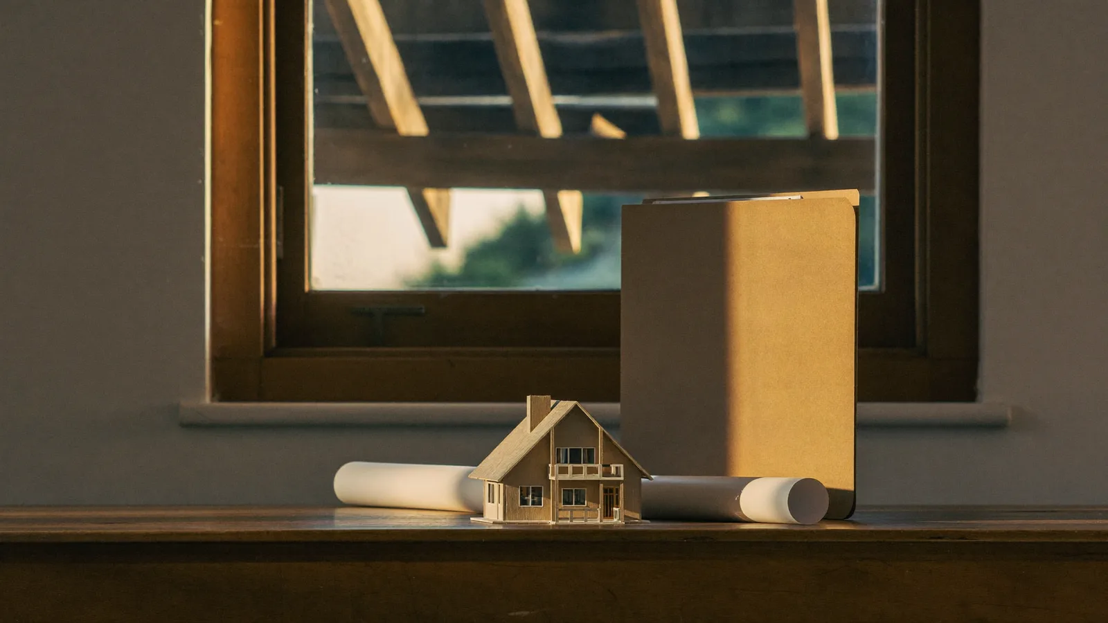 Maquette de maison en bois, plan roulé et chemise cartonnée sur un appui de fenêtre, charpente ancienne en arrière-plan dans une lumière dorée